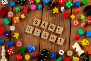 "Game Night" spelled out in wooden letter tiles. Surrounded by dice, cards, and other game pieces on a wooden background